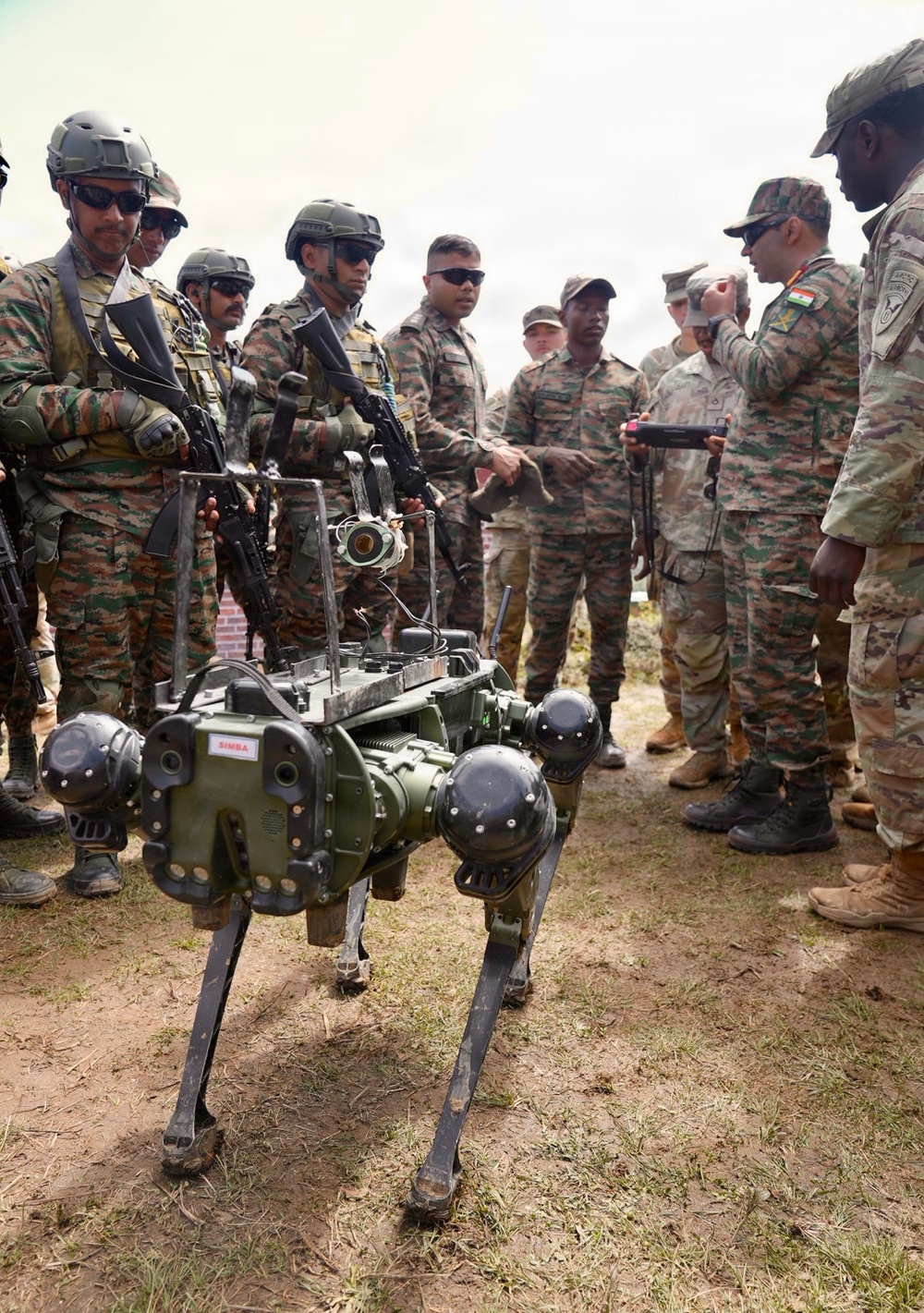 A group of military personnel from what appears to be Indian Army and US Army forces are gathered around a quadruped military robot labeled 'SIMBA'. The robot is a four-legged autonomous ground vehicle painted in olive/dark green, equipped with sensors and cameras. A soldier appears to be demonstrating or operating the robot using a handheld controller.