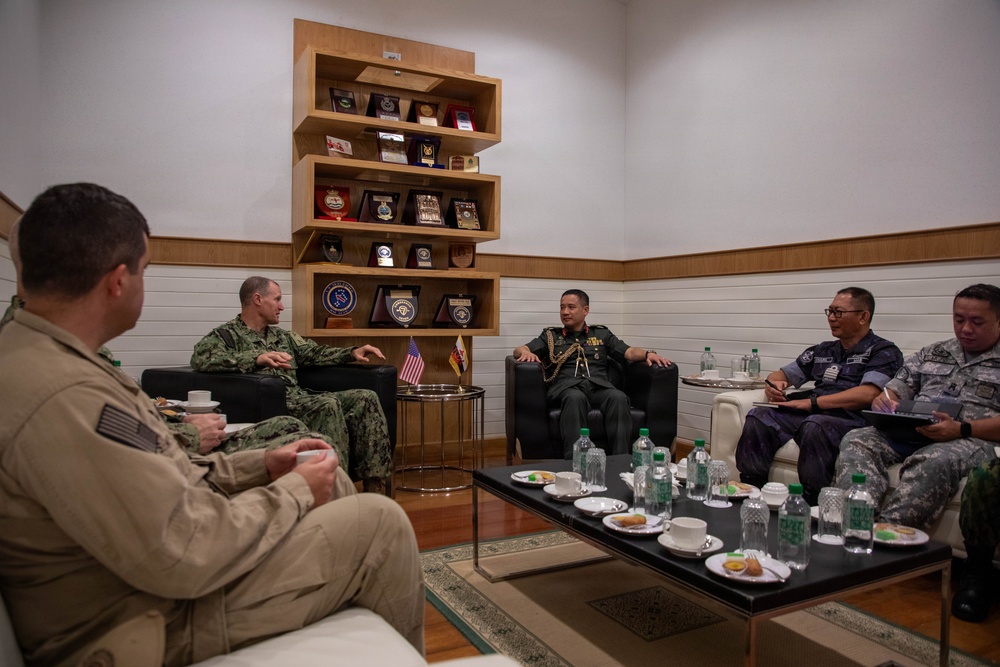 A formal military meeting is taking place in an official reception room between personnel wearing U.S. Navy uniforms and individuals in what appear to be Southeast Asian military/police uniforms. The setting includes a display shelf with military plaques and commemorative items, and a small American flag is visible on a side table. The group is seated in a circular arrangement around a coffee table with water bottles, cups, and light refreshments.