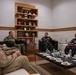 A formal military meeting is taking place in an official reception room between personnel wearing U.S. Navy uniforms and individuals in what appear to be Southeast Asian military/police uniforms. The setting includes a display shelf with military plaques and commemorative items, and a small American flag is visible on a side table. The group is seated in a circular arrangement around a coffee table with water bottles, cups, and light refreshments.