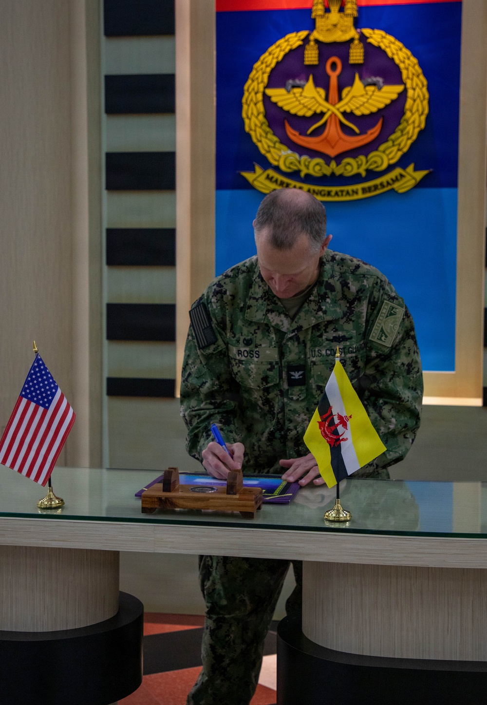 A U.S. Coast Guard officer named Ross is signing a document at a glass-topped table during what appears to be an official bilateral meeting or ceremony. The setting is a formal military headquarters, as indicated by the 'Markas Angkatan Bersama' (Joint Forces Headquarters) emblem visible on the wall behind him. Both the United States and Brunei flags are displayed on the table, indicating a U.S.-Brunei military engagement.