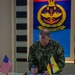 A U.S. Coast Guard officer named Ross is signing a document at a glass-topped table during what appears to be an official bilateral meeting or ceremony. The setting is a formal military headquarters, as indicated by the 'Markas Angkatan Bersama' (Joint Forces Headquarters) emblem visible on the wall behind him. Both the United States and Brunei flags are displayed on the table, indicating a U.S.-Brunei military engagement.