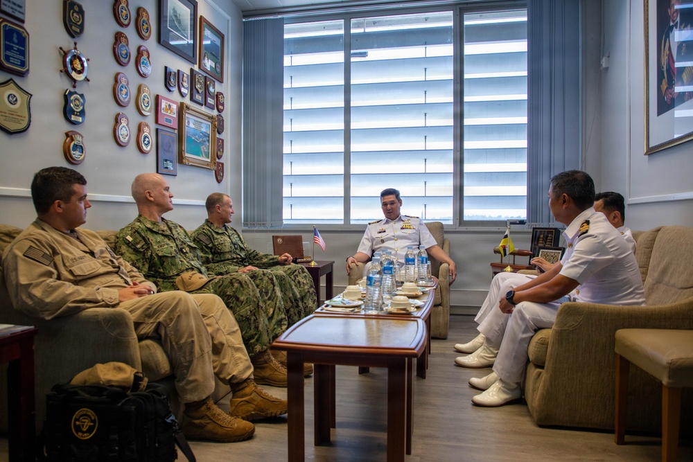 A formal military meeting is taking place in an office setting between personnel in U.S. military uniforms (camouflage and Army Combat Uniform) and personnel in white naval dress uniforms, likely from an Asian navy. Both delegations are seated on sofas around a coffee table with water bottles and cups. The office walls display numerous military plaques, crests, and framed photographs, and both a U.S. flag and what appears to be a Brunei flag are visible on the desk.