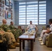 A formal military meeting is taking place in an office setting between personnel in U.S. military uniforms (camouflage and Army Combat Uniform) and personnel in white naval dress uniforms, likely from an Asian navy. Both delegations are seated on sofas around a coffee table with water bottles and cups. The office walls display numerous military plaques, crests, and framed photographs, and both a U.S. flag and what appears to be a Brunei flag are visible on the desk.