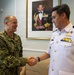 Two military officers are shaking hands in what appears to be an official meeting room. One officer wears U.S. Navy digital camouflage (NWU) with the name tape 'ROSS,' while the other wears a white naval dress uniform with multiple ribbons and insignia. A formal portrait of a dignitary in military dress hangs on the wall behind them.