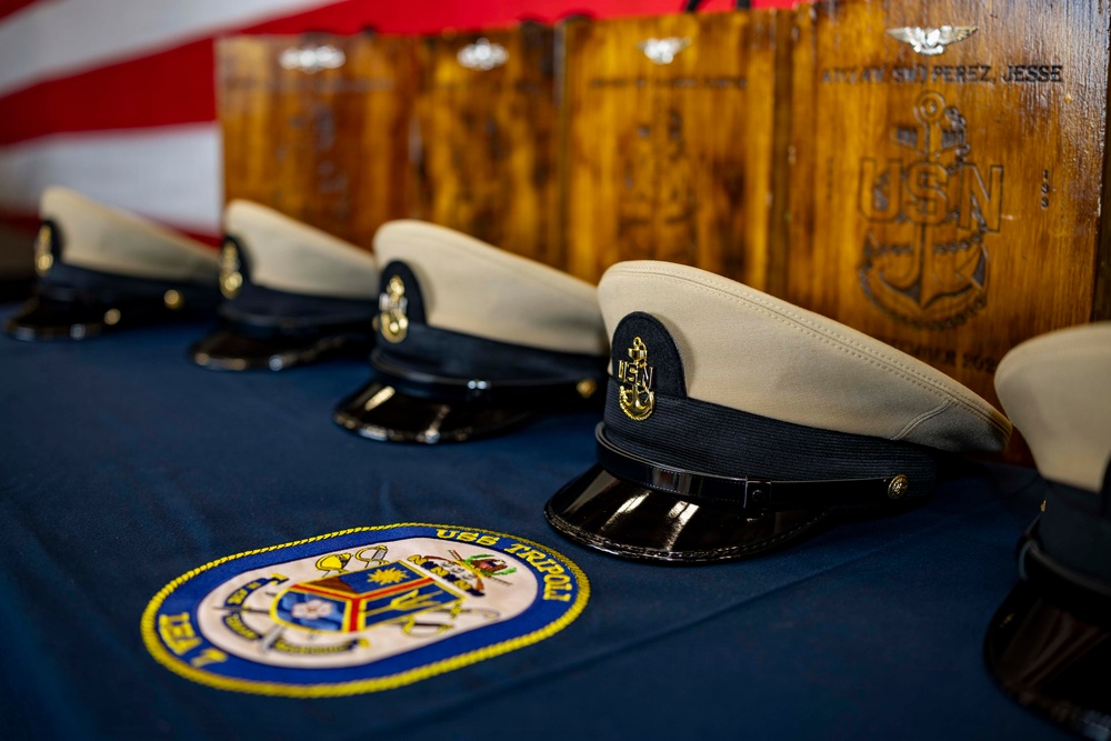 The image shows a ceremonial display of U.S. Navy Chief Petty Officer covers (hats) arranged on a blue tablecloth bearing the USS Tripoli ship patch. Behind the hats are wooden plaques engraved with the USN anchor insignia and what appears to be a sailor's name 'Attanasio Perez, Jesse'. An American flag is partially visible in the upper left background, suggesting this is a military promotion or advancement ceremony.