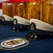 The image shows a ceremonial display of U.S. Navy Chief Petty Officer covers (hats) arranged on a blue tablecloth bearing the USS Tripoli ship patch. Behind the hats are wooden plaques engraved with the USN anchor insignia and what appears to be a sailor's name 'Attanasio Perez, Jesse'. An American flag is partially visible in the upper left background, suggesting this is a military promotion or advancement ceremony.