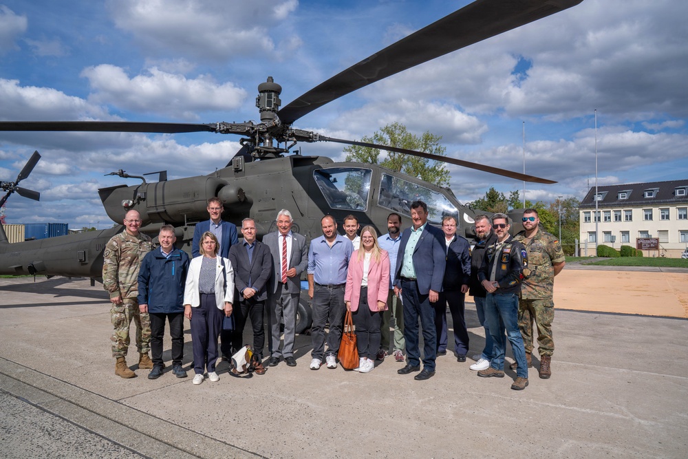 A group of approximately 15 individuals, including military personnel and civilians in business attire, pose for a group photo in front of a military attack helicopter, likely an AH-64 Apache. The setting appears to be a military installation or base, with institutional buildings visible in the background. The mixed group suggests an official visit, delegation, or diplomatic engagement at a military facility.