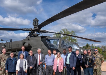 A group of approximately 15 individuals, including military personnel and civilians in business attire, pose for a group photo in front of a military attack helicopter, likely an AH-64 Apache. The setting appears to be a military installation or base, with institutional buildings visible in the background. The mixed group suggests an official visit, delegation, or diplomatic engagement at a military facility.