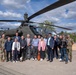 A group of approximately 15 individuals, including military personnel and civilians in business attire, pose for a group photo in front of a military attack helicopter, likely an AH-64 Apache. The setting appears to be a military installation or base, with institutional buildings visible in the background. The mixed group suggests an official visit, delegation, or diplomatic engagement at a military facility.