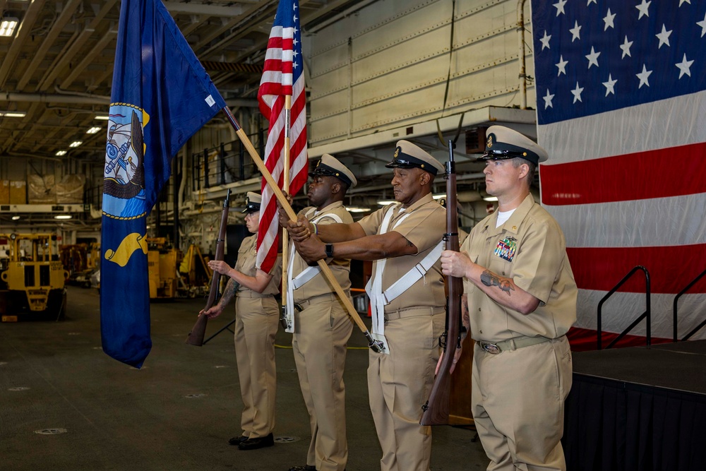 A U.S. Navy color guard is performing a formal ceremony, likely a change of command or commissioning event, inside what appears to be a ship's hangar bay. Four sailors in khaki service dress uniforms are presenting the colors, carrying the American flag, a Navy unit flag, and ceremonial rifles. A large American flag is displayed as a backdrop on the right side of the frame.