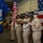 USS Tripoli Chief Pinning A U.S. Navy color guard is performing a formal ceremony, likely a change of command or commissioning event, inside what appears to be a ship's hangar bay. Four sailors in khaki service dress uniforms are presenting the colors, carrying the American flag, a Navy unit flag, and ceremonial rifles. A large American flag is displayed as a backdrop on the right side of the frame.