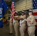 A U.S. Navy color guard is performing a formal ceremony, likely a change of command or commissioning event, inside what appears to be a ship's hangar bay. Four sailors in khaki service dress uniforms are presenting the colors, carrying the American flag, a Navy unit flag, and ceremonial rifles. A large American flag is displayed as a backdrop on the right side of the frame.