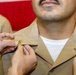 A military pinning ceremony is taking place, with two sets of hands attaching gold insignia pins to the collar of a service member wearing a tan/khaki military uniform. The recipient appears to be a U.S. Navy or Marine Corps member based on the uniform color and style. A second uniformed individual with ribbons and medals is visible in the background.