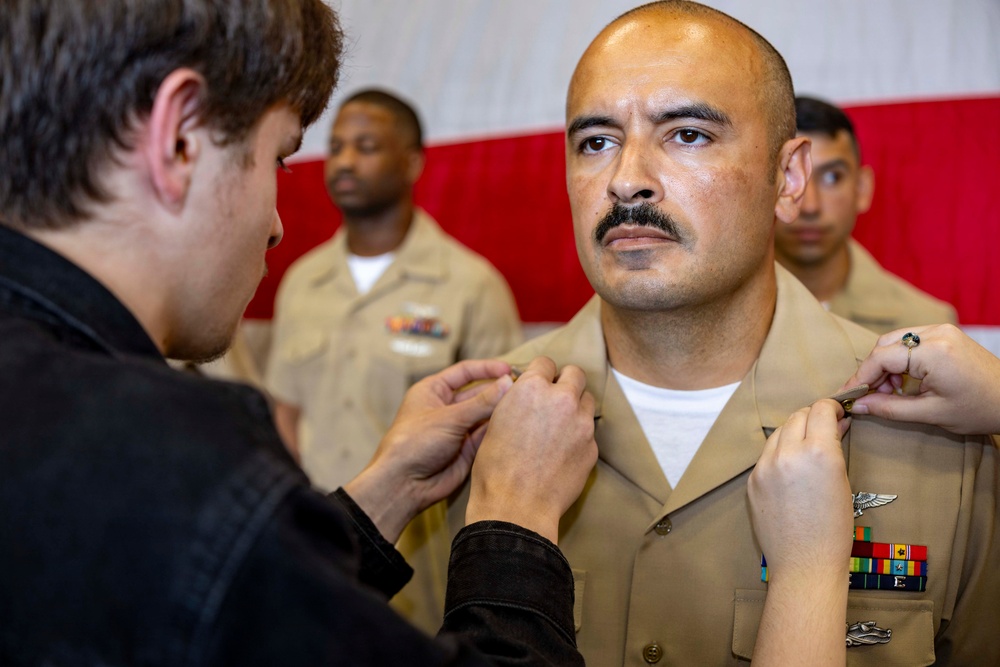 A U.S. Navy sailor in khaki service uniform is having insignia or rank devices pinned onto his collar by two individuals during what appears to be a promotion or advancement ceremony. The sailor stands at attention with a composed expression while others in similar uniforms are visible in the background. A red and white backdrop, likely a flag or banner, is visible behind the group.