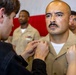 A U.S. Navy sailor in khaki service uniform is having insignia or rank devices pinned onto his collar by two individuals during what appears to be a promotion or advancement ceremony. The sailor stands at attention with a composed expression while others in similar uniforms are visible in the background. A red and white backdrop, likely a flag or banner, is visible behind the group.