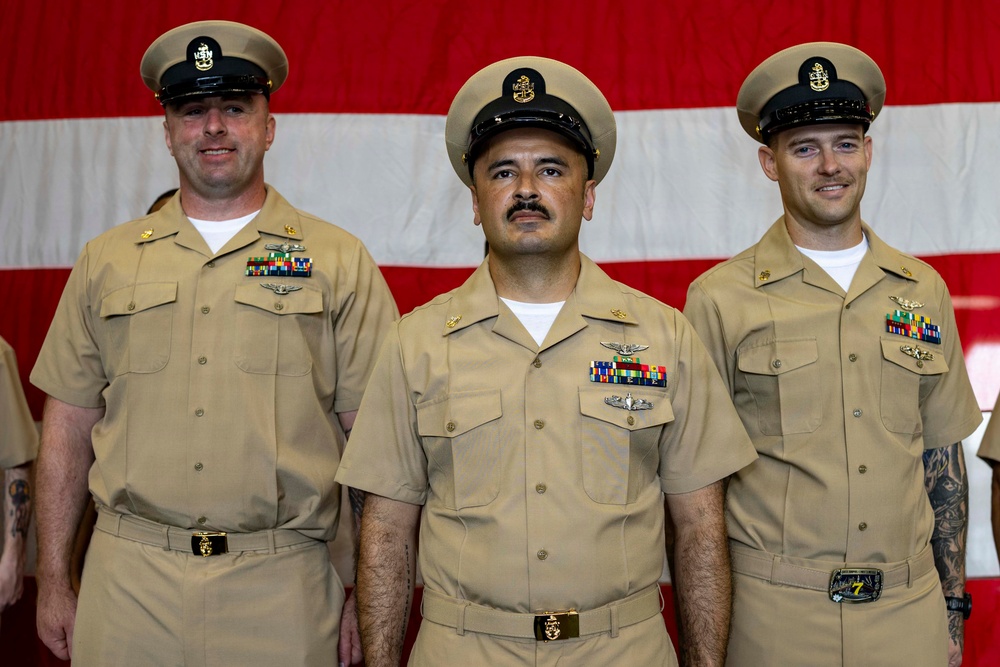 Three U.S. Navy enlisted personnel in Service Khaki uniforms are standing in formation in front of a large American flag backdrop. All three wear Chief Petty Officer or Senior Chief Petty Officer covers and display multiple service ribbons, warfare qualification badges, and rank insignia. The setting appears to be a formal military ceremony or official photo.