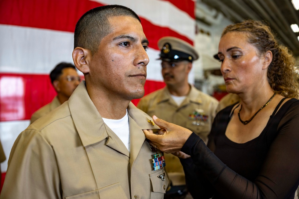 A military promotion or award ceremony is taking place, likely in a naval or Marine Corps setting. A woman in civilian clothing is pinning an insignia or device onto the collar of a service member in a tan/khaki uniform. An American flag and additional uniformed personnel are visible in the background.