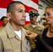 A military promotion or award ceremony is taking place, likely in a naval or Marine Corps setting. A woman in civilian clothing is pinning an insignia or device onto the collar of a service member in a tan/khaki uniform. An American flag and additional uniformed personnel are visible in the background.
