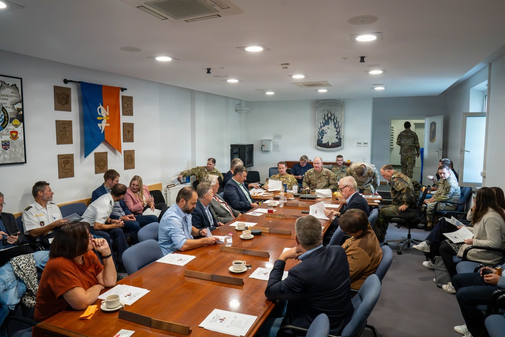 A formal meeting or briefing is taking place in what appears to be a military conference room, with a mix of uniformed military personnel and civilians seated around a large wooden table. The room is decorated with military unit plaques, insignia, and a flag bearing what appears to be a NATO-affiliated emblem. Personnel are engaged in reviewing documents and conducting discussions.