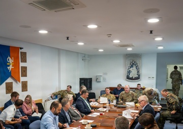 A formal meeting or briefing is taking place in what appears to be a military conference room, with a mix of uniformed military personnel and civilians seated around a large wooden table. The room is decorated with military unit plaques, insignia, and a flag bearing what appears to be a NATO-affiliated emblem. Personnel are engaged in reviewing documents and conducting discussions.