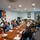 U.S. Army, Bundeswehr, and Local Mayors Align for Noise Abatement Meeting and Chinook Helicopter Flight A formal meeting or briefing is taking place in what appears to be a military conference room, with a mix of uniformed military personnel and civilians seated around a large wooden table. The room is decorated with military unit plaques, insignia, and a flag bearing what appears to be a NATO-affiliated emblem. Personnel are engaged in reviewing documents and conducting discussions.