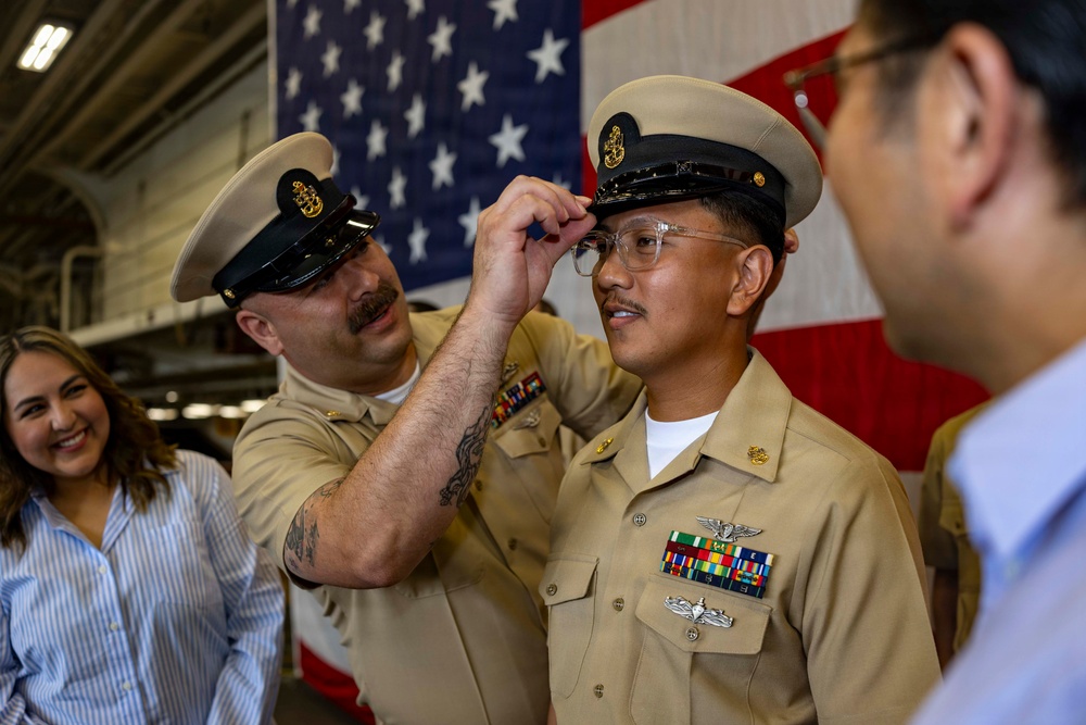 A U.S. Navy promotion or advancement ceremony is taking place in what appears to be a hangar bay, with an American flag displayed prominently in the background. One Navy chief petty officer is placing a combination cover (cap) on another sailor who has just been advanced, likely to the rank of Chief Petty Officer. Onlookers, including a smiling civilian woman, observe the moment.