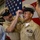 USS Tripoli Chief Pinning A U.S. Navy promotion or advancement ceremony is taking place in what appears to be a hangar bay, with an American flag displayed prominently in the background. One Navy chief petty officer is placing a combination cover (cap) on another sailor who has just been advanced, likely to the rank of Chief Petty Officer. Onlookers, including a smiling civilian woman, observe the moment.