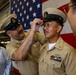 A U.S. Navy promotion or advancement ceremony is taking place in what appears to be a hangar bay, with an American flag displayed prominently in the background. One Navy chief petty officer is placing a combination cover (cap) on another sailor who has just been advanced, likely to the rank of Chief Petty Officer. Onlookers, including a smiling civilian woman, observe the moment.