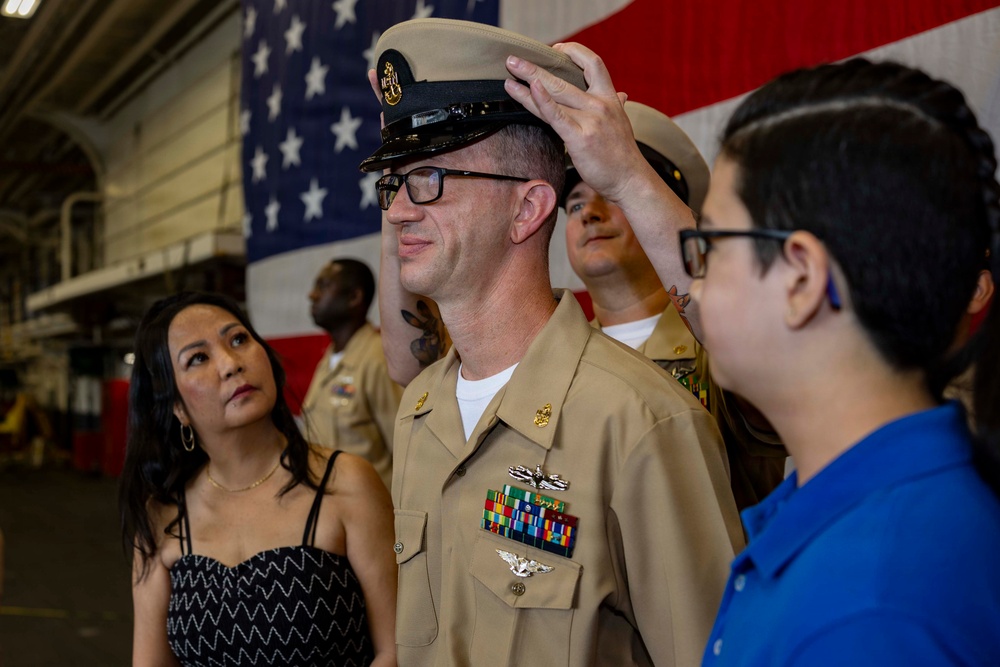 A U.S. Navy chief petty officer promotion ceremony is taking place in what appears to be a ship's hangar bay. A young person, likely a family member, is placing the chief's combination cover (hat) on the newly promoted sailor's head, a traditional part of the CPO pinning/frocking ceremony. A woman in a black dress, presumably a family member, watches the moment alongside other personnel in the background.