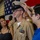 USS Tripoli Chief Pinning A U.S. Navy chief petty officer promotion ceremony is taking place in what appears to be a ship's hangar bay. A young person, likely a family member, is placing the chief's combination cover (hat) on the newly promoted sailor's head, a traditional part of the CPO pinning/frocking ceremony. A woman in a black dress, presumably a family member, watches the moment alongside other personnel in the background.
