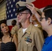 A U.S. Navy chief petty officer promotion ceremony is taking place in what appears to be a ship's hangar bay. A young person, likely a family member, is placing the chief's combination cover (hat) on the newly promoted sailor's head, a traditional part of the CPO pinning/frocking ceremony. A woman in a black dress, presumably a family member, watches the moment alongside other personnel in the background.