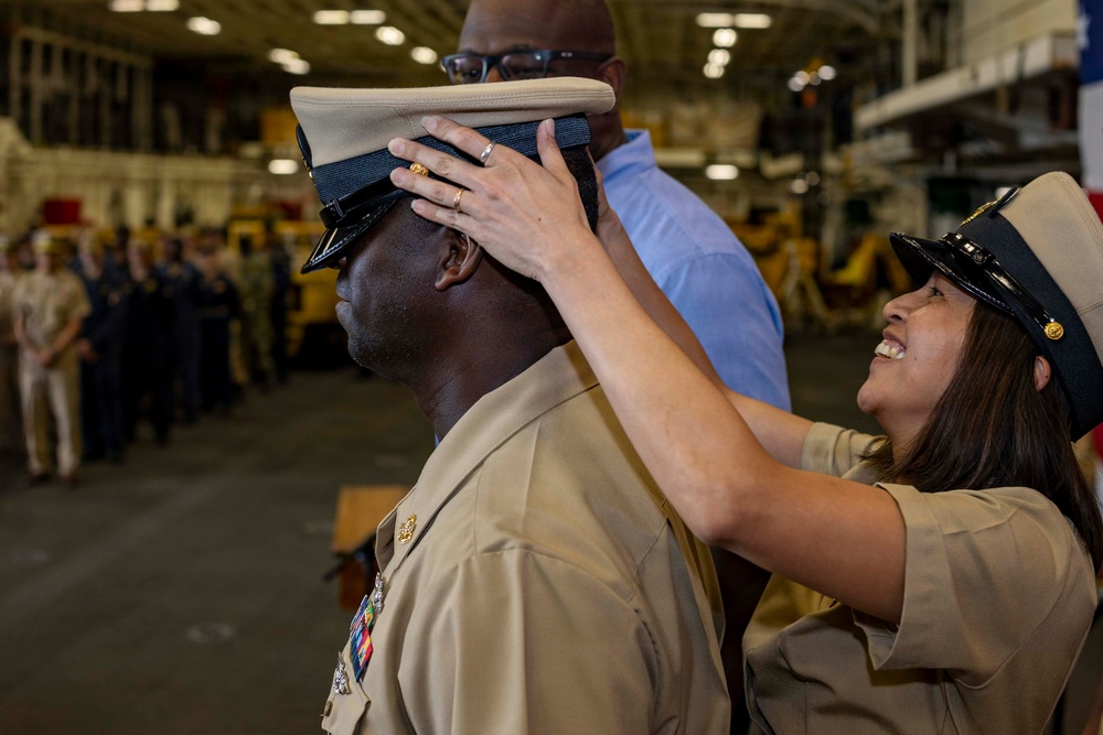 A military promotion or advancement ceremony is taking place in what appears to be a ship's hangar bay. A woman in a tan/khaki Navy uniform is placing a chief petty officer's combination cover (hat) onto a male service member's head, a traditional part of the CPO pinning/advancement ceremony. Rows of uniformed personnel are visible in the background observing the ceremony.