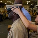 A military promotion or advancement ceremony is taking place in what appears to be a ship's hangar bay. A woman in a tan/khaki Navy uniform is placing a chief petty officer's combination cover (hat) onto a male service member's head, a traditional part of the CPO pinning/advancement ceremony. Rows of uniformed personnel are visible in the background observing the ceremony.