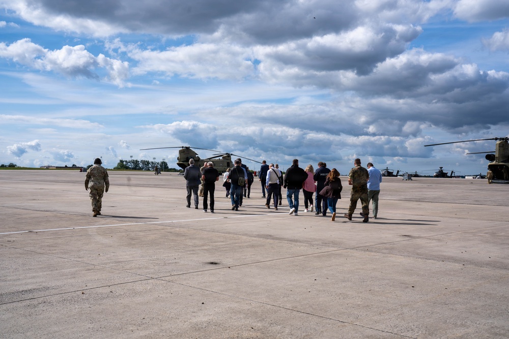 A group of civilians and military personnel are walking across a military airfield tarmac toward large tandem-rotor helicopters, likely CH-47 Chinooks. At least two soldiers in camouflage uniforms are escorting the civilian group, suggesting an official visit, tour, or briefing. Additional helicopters, possibly Apache attack helicopters, are visible on the right side of the frame.