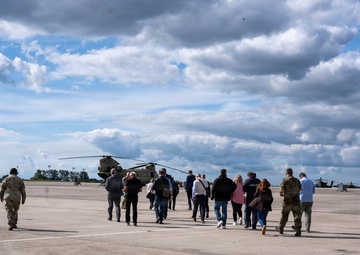 A group of civilians and military personnel are walking across a military airfield tarmac toward large tandem-rotor helicopters, likely CH-47 Chinooks. At least two soldiers in camouflage uniforms are escorting the civilian group, suggesting an official visit, tour, or briefing. Additional helicopters, possibly Apache attack helicopters, are visible on the right side of the frame.