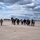 U.S. Army, Bundeswehr, and Local Mayors Align for Noise Abatement Meeting and Chinook Helicopter Flight A group of civilians and military personnel are walking across a military airfield tarmac toward large tandem-rotor helicopters, likely CH-47 Chinooks. At least two soldiers in camouflage uniforms are escorting the civilian group, suggesting an official visit, tour, or briefing. Additional helicopters, possibly Apache attack helicopters, are visible on the right side of the frame.