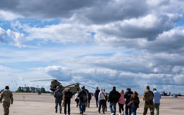 A group of civilians and military personnel are walking across a military airfield tarmac toward large tandem-rotor helicopters, likely CH-47 Chinooks. At least two soldiers in camouflage uniforms are escorting the civilian group, suggesting an official visit, tour, or briefing. Additional helicopters, possibly Apache attack helicopters, are visible on the right side of the frame.