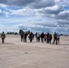 A group of civilians and military personnel are walking across a military airfield tarmac toward large tandem-rotor helicopters, likely CH-47 Chinooks. At least two soldiers in camouflage uniforms are escorting the civilian group, suggesting an official visit, tour, or briefing. Additional helicopters, possibly Apache attack helicopters, are visible on the right side of the frame.