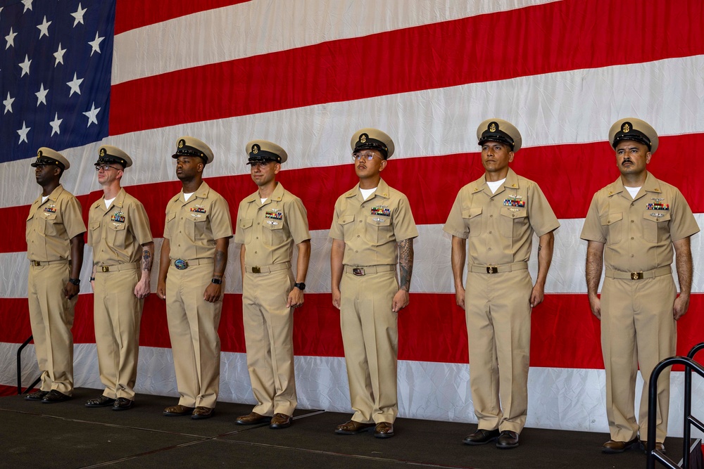 Seven U.S. Navy personnel in khaki service uniforms stand at attention in a formal line on a stage or platform. A large American flag serves as the backdrop, suggesting an official military ceremony such as a promotion, change of command, or chief petty officer pinning event. The personnel display various service ribbons and rank insignia on their uniforms.