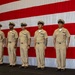 Seven U.S. Navy personnel in khaki service uniforms stand at attention in a formal line on a stage or platform. A large American flag serves as the backdrop, suggesting an official military ceremony such as a promotion, change of command, or chief petty officer pinning event. The personnel display various service ribbons and rank insignia on their uniforms.
