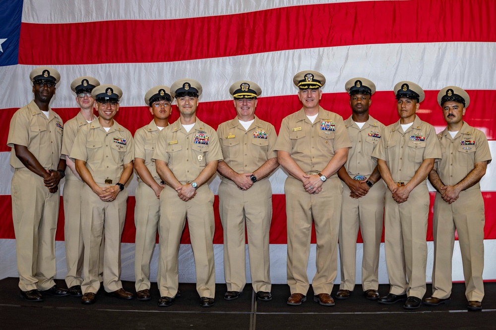A formal group portrait of nine United States Navy personnel in khaki service uniforms standing in front of a large American flag backdrop. The group includes personnel of varying ranks, identifiable by their cover devices, collar insignia, and ribbon/medal arrangements. This appears to be an official military ceremony or recognition event.