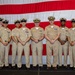 A formal group portrait of nine United States Navy personnel in khaki service uniforms standing in front of a large American flag backdrop. The group includes personnel of varying ranks, identifiable by their cover devices, collar insignia, and ribbon/medal arrangements. This appears to be an official military ceremony or recognition event.