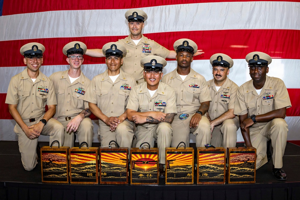 A group of eight U.S. Navy Chief Petty Officers pose for a formal group photograph in front of an American flag backdrop. They are dressed in khaki service uniforms with combination covers (white-topped caps) and display various ribbons and warfare devices. In front of them are seven decorative wooden boxes featuring Japanese-style artwork with kanji characters and sunrise/Mount Fuji imagery.