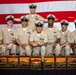A group of eight U.S. Navy Chief Petty Officers pose for a formal group photograph in front of an American flag backdrop. They are dressed in khaki service uniforms with combination covers (white-topped caps) and display various ribbons and warfare devices. In front of them are seven decorative wooden boxes featuring Japanese-style artwork with kanji characters and sunrise/Mount Fuji imagery.