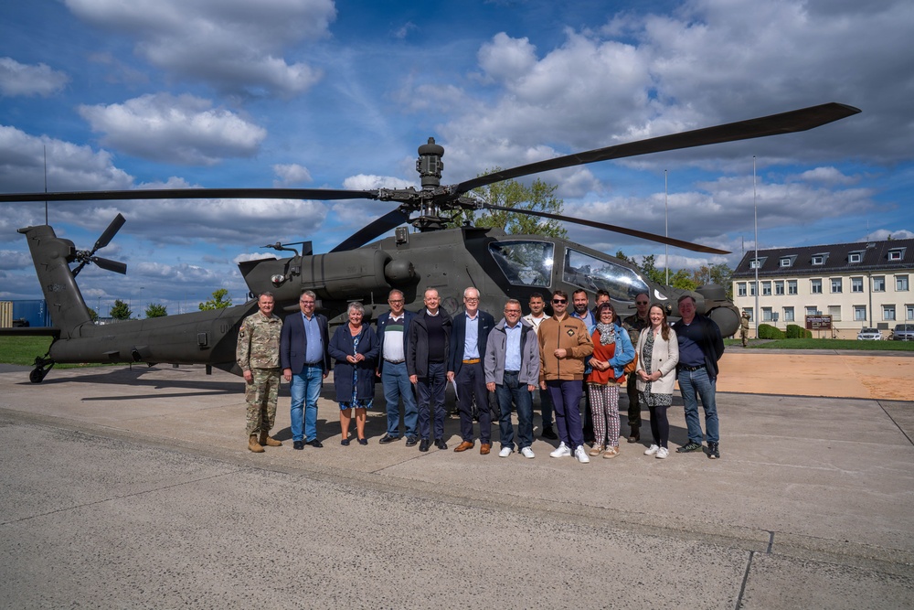 A group of approximately 14 civilians and one military personnel in uniform are posing for a group photograph in front of a military attack helicopter, likely an AH-64 Apache, on a paved tarmac area. The setting appears to be a military installation or base, evidenced by the institutional building visible in the background. The occasion appears to be an official visit or delegation tour of the military facility.