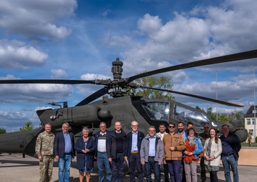 A group of approximately 14 civilians and one military personnel in uniform are posing for a group photograph in front of a military attack helicopter, likely an AH-64 Apache, on a paved tarmac area. The setting appears to be a military installation or base, evidenced by the institutional building visible in the background. The occasion appears to be an official visit or delegation tour of the military facility.