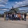 U.S. Army, Bundeswehr, and Local Mayors Align for Noise Abatement Meeting and Chinook Helicopter Flight A group of approximately 14 civilians and one military personnel in uniform are posing for a group photograph in front of a military attack helicopter, likely an AH-64 Apache, on a paved tarmac area. The setting appears to be a military installation or base, evidenced by the institutional building visible in the background. The occasion appears to be an official visit or delegation tour of the military facility.