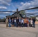 A group of approximately 14 civilians and one military personnel in uniform are posing for a group photograph in front of a military attack helicopter, likely an AH-64 Apache, on a paved tarmac area. The setting appears to be a military installation or base, evidenced by the institutional building visible in the background. The occasion appears to be an official visit or delegation tour of the military facility.