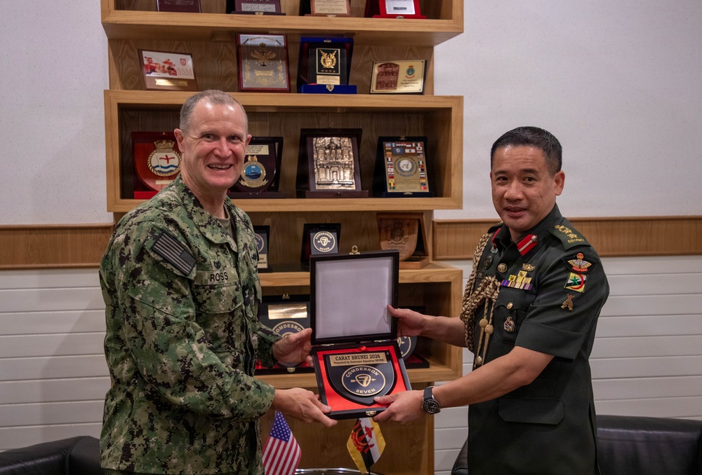Two military officers, one in U.S. Navy digital camouflage uniform and one in a dark green uniform with ceremonial decorations, are jointly presenting a commemorative plaque or award. The plaque reads 'CARAT BRUNEI 2026' and 'COMDESRON SEVEN', indicating a bilateral military exercise exchange ceremony. Both men are smiling, suggesting a formal but cordial diplomatic military engagement.