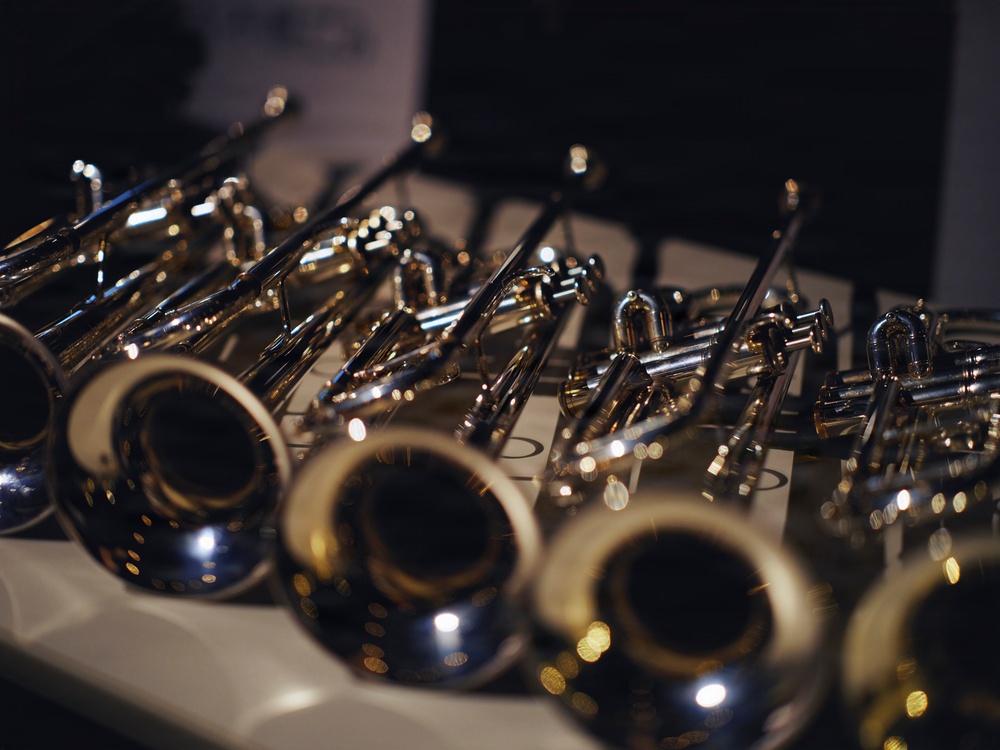 The image shows a collection of brass wind instruments, specifically trumpets, arranged closely together on a flat surface. The instruments display polished silver and gold finishes with characteristic bell openings facing the camera in the foreground. The shallow depth of field creates a bokeh effect, keeping only the nearest instruments in sharp focus.